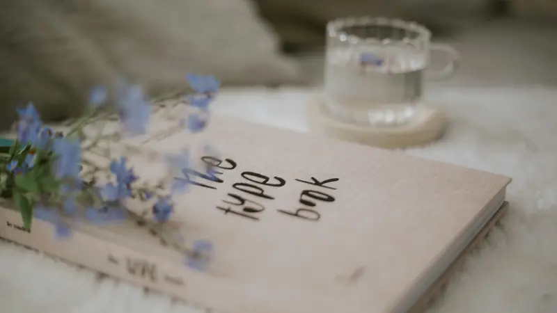 Un carnet posé sur un plaid, à côté d'un verre d'eau et de fleurs bleues.