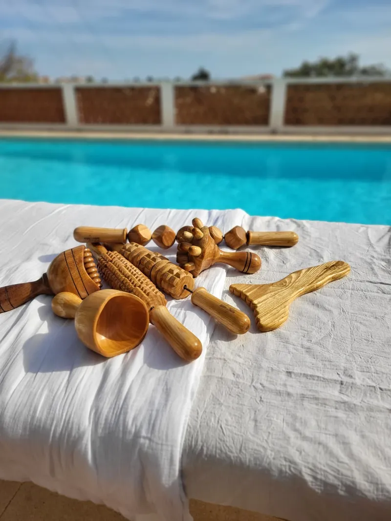 Les instruments de madérothérapie en bois de Madero Sud — rouleaux, coupe, planche — posés sur un drap blanc, au bord d'une piscine.